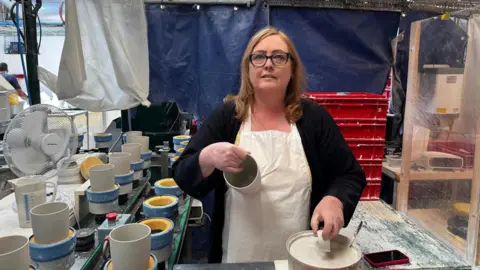 Sharon has shoulder-length brown hair and is wearing glasses. She has a white apron on and is holding a cup. Around her are other mugs waiting to have handles put on them