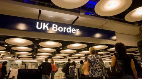 In Pictures via Getty Images Images UK airport passport control. There is a blue overhead sign with UK border written on it in white. People are queuing to get their passports checked.