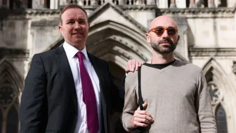 Bloomberg via Getty Images Two men, one in a suit and purple tie, the other in glasses and a brown jumper, stand outside a grand building