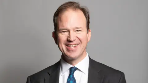 UK Parliament A portrait photograph of MP Jesse Norman. He is wearing a grey suit jacket and white shirt with a blue spotty tie. He is stood against a grey backdrop.