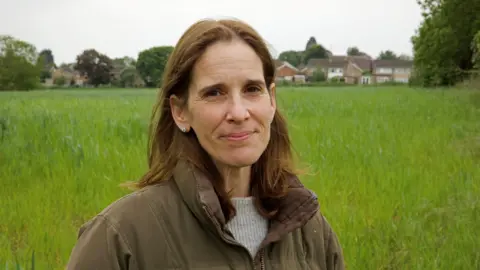 Joe Crowley / BBC A head-and-shoulders portrait of Kate Pryke, a white woman with long brown hair who is wearing a brown padded jacket over a ribbed grey jumper, standing in a green field, with houses in the distance