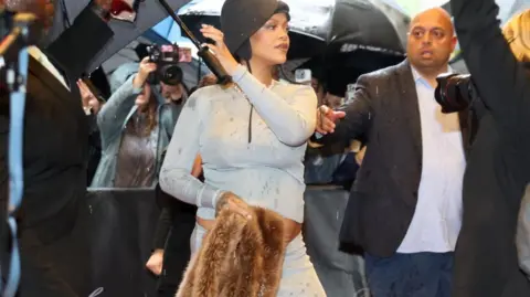 Reuters Rihanna is pictured wearing a pale blue outfit and holding a piece of clothing obscuring her midriff. She is pictured under an umbrella at the Carlyle Hotel before attending the Met Gala.