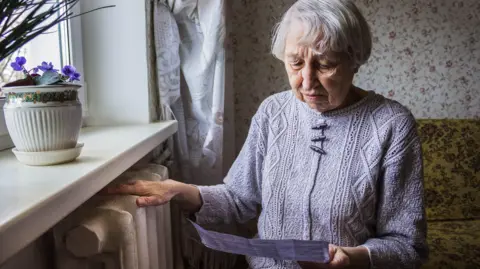 Age UK An elderly lady in her front room looks anxiously at a paper bill in her hand, with her other hand resting on a radiator. She has silver hair and is wearing a knitted jumper. 