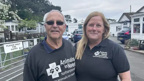 Kate Bradbrook/BBC A man and woman wearing branded T-shirts stand in front of gates at the entrance to an animal sanctuary.