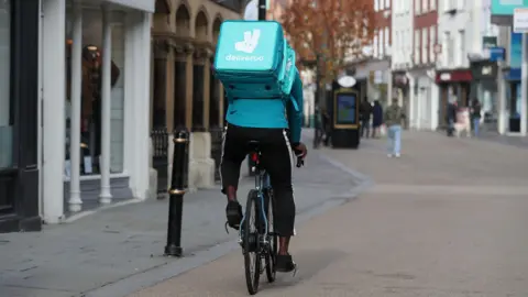 PA Deliveroo rider on bike wearing black trousers, bright blue top, and bright blue square Deliveroo bag on his back saying the firm's name and with its rabbit logo on it