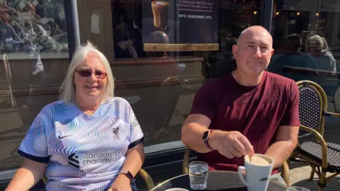 Susan and Wayne Thirwell sit at an outdoor table at a coffee shop both smiling at the camera