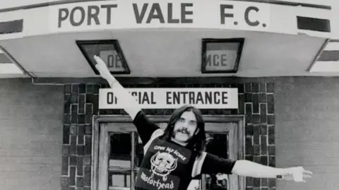 IFK Legacy CIC A black and white photograph of Motörhead frontman Lemmy, a man with long dark hair and a dark moustache, holding his arms out like a plane underneath a sign reading