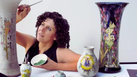 Moorcroft A woman can be seen against a white backdrop, painting tall pieces of pottery. She is wearing a black sleeveless top and is holding a bowl of green paint.