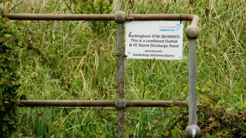 Joe Crowley / BBC The bank of the Great Ouse river, with long grass and bushes growing around metal railings with signage from Anglian Water, which says