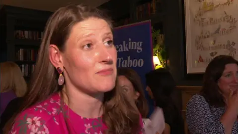 BBC Theo Clarke stands in front of a banner adverting her book. She has shoulder-length dark hair and is wearing a pink dress and has pink earrings.