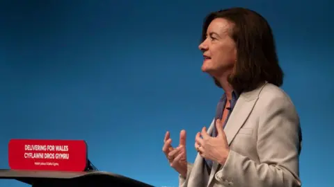 Getty Images Eluned Morgan stood at a podium delivering a speech at a party conference. She is wearing a beige blazer and is gesturing with her hands.