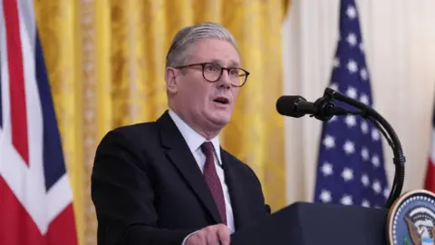 EPA Sir Keir Starmer in a suit and tie stands at a podium in the White House with the presidential seal, flanked by British and American flags.