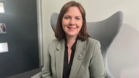 BBC Woman with short brown hair in green linen jacket with black blouse and silver broach sits in grey armchair