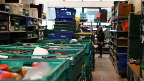 Cambridge City Foodbank In the centre of a rectangle shaped room are stacks of green plastic trays. These are the food parcels given to foodbank users. At the sides of the room are shelves with more trays on. A person stands in the background holding a piece of paper while looking at the food parcels.