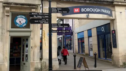 A shot of part of Chippenham centre - the entrance to the Borough Parade Shopping area with a large signpost in front and the pawesome pet shop to the left.