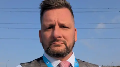 Facebook Joel Tetlow has short brown hair and a beard. He is wearing a shirt, waistcoat and pink tie, with the top of a Reform UK lanyard just visible.