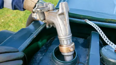 Getty Images A silver pump is unloading a home heating oil delivery into a green plastic tank. You can see a person holding the pump. They are wearing a grey rubber glove with a blue and white sleeve.