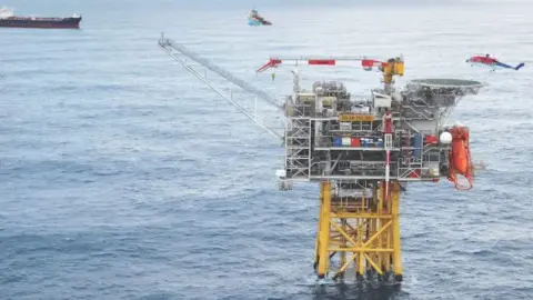 Harbour Energy Oil and gas platform with yellow legs in the sea, a blue, red and white offshore helicopter hovers overhead, and vessels in the background.