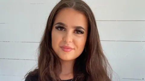 Victoria Calvert Labubu fan Victoria Calvert who has long brown hair and pink lipstick smiles at the camera next to a white wall