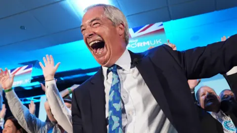 PA Media Nigel Farage - a man with silver hair and dressed in a suit with blue tie - laughs happily onstage while surrounded by supporters