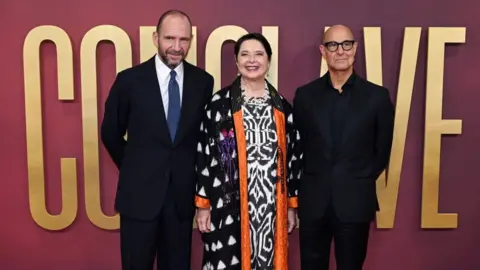 Getty Images Ralph Fiennes, Isabella Rossellini and Stanley Tucci stand in front of a purple wall with large gold text that says