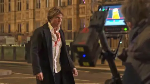 Paramount Pictures Tom Cruise appears pensive in front of a camera outside the Palace of Westminster. He is wearing a black coat, with a white shirt, slightly open and smeared with red liquid