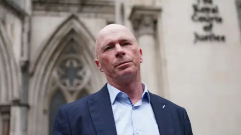 PA Media Raymond Connolly standing outside the Royal Courts of Justice in London. He is looking down the barrel of the camera and is wearing a light blue, open-necked shirt with a dark blue jacket. He has a bald head and his expression is serious. The Gothic arched windows of the grey stone court building can be seen over his right shoulder, while the words