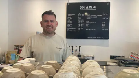 BBC Bradley Buechel wears a grey t-shirt and looks into the camera from behind his professional coffee machine. A price list is behind him on the wall and there are coffee cups lined up on the top of the machine.