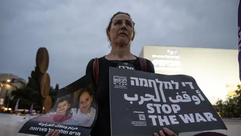 Getty Images A woman stands in protest holding up two signs - one showing the image of two Gazan children killed in the war and another that says in Hebrew, Arabic and English: 