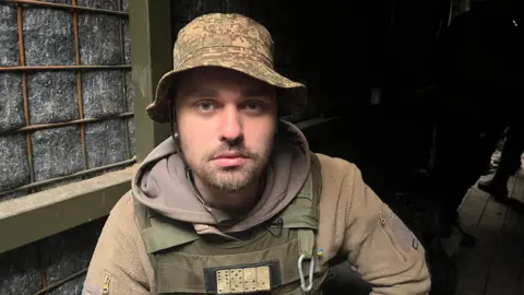 BBC A Ukrainian soldier wears camouflage and looks directly into the camera. He has a brown hoody underneath a dark green bulletproof vest and wears a bucket hat. He has a beard and moustache and wears a forlorn expression.