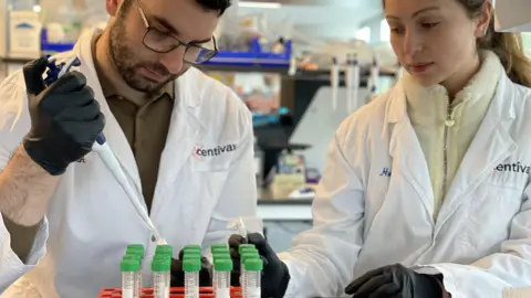 Jacob Glanville Two scientists wearing white lab coats and black gloves are pipetting into small vials in a laboratory